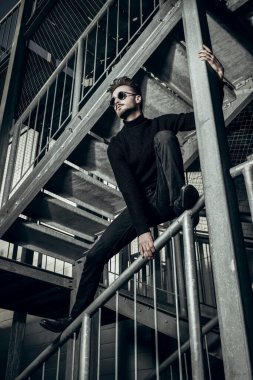 Siyah elbiseli ve siyah güneş gözlüklü yakışıklı bir adamın portresi. Metal endüstriyel bir merdivende poz veriyor. Şehirli grunge stili. Erkek modası.