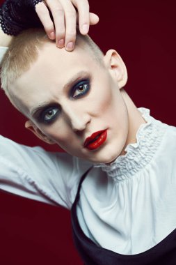 Kadın makyajlı ve kadın kıyafetleriyle stüdyoda ifade ile poz veren genç bir adamın tam boy portresi..