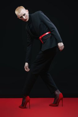 Kadın makyajlı ve kadın kıyafetleriyle stüdyoda ifade ile poz veren genç bir adamın tam boy portresi..