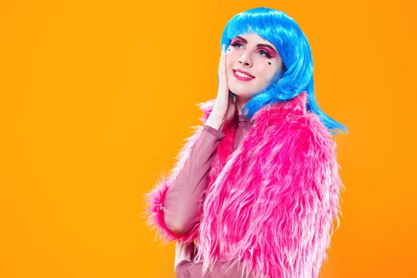 Portrait of an attractive party girl with bright pink makeup and blue wig wearing pink fur coat on a yellow background. Make-up and cosmetics, hairstyle. Fashion girl. Copy space.