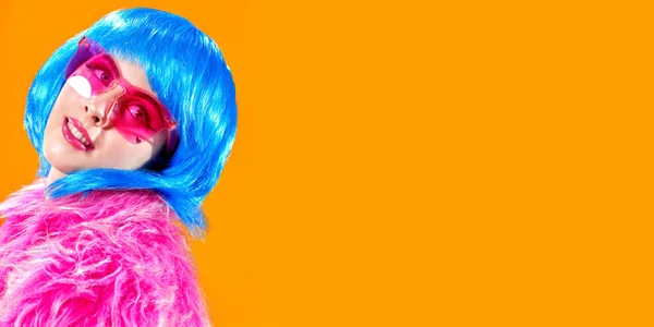 Portrait of an attractive party girl with bright pink makeup and blue wig wearing pink fur coat and sunglasses on a yellow background. Make-up and cosmetics, hairstyle. Fashion girl. Copy space.