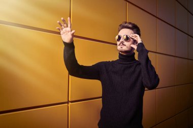 Şehir tarzı. Siyah kazaklı ve yuvarlak aynalı genç bir adamın portresi sarı sanayi duvarının yanındaki sokakta duran bir şeye dokunmak için elini kaldırdı. Erkek modası. Boşluğu kopyala.