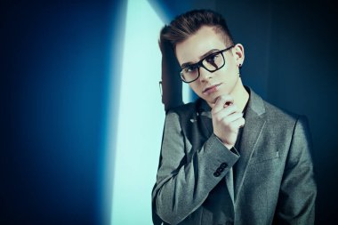 Klasik takım elbiseli ve gözlüklü yakışıklı genç adam stüdyoda mavi ışıkta beyaz duvarın yanında poz veriyor. Eğitim, eğitim. Erkek güzelliği, moda. 