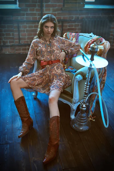 Bohemian style. Fashion shot of a beautiful young woman in boho style dress and jewelry posing in a vintage armchair. Full length portrait.