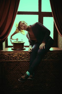 Şık takım elbiseli yakışıklı genç adam, şık bir iç mekanda pencere eşiğinde oturuyor. Erkek güzelliği, moda. 