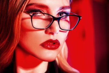 Kırmızı ışıkta gözlük takan kendine güvenen bir kadının yakın portresi. Güzellik, makyaj, tarz.