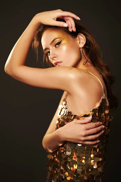 Luxurious lifestyle. Portrait of a glamorous young woman with golden make-up in golden dress on a dark grey background. Party style. Copy space.