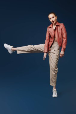 Yeni bahar stili. Koyu mavi bir arka planda hareket halinde deri ceketle poz veren sevimli, modern bir kız modelin portresi. Güzellik, moda. 