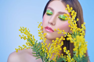 Bahar güzeli. Mavi arka planda parlak makyajlı ve sarı mimoza çiçekli hassas bir genç kadının portresi. Makyaj ve makyaj malzemeleri. Boşluğu kopyala.