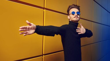 Şehir tarzı. Siyah kazaklı şık bir gencin portresi ve sarı sanayi duvarının yanındaki bir sokakta poz veren yuvarlak aynalı güneş gözlüğü. Erkek modası. Boşluğu kopyala.