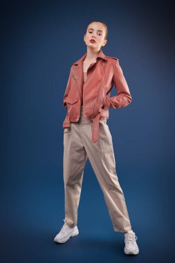 Yeni bahar stili. Koyu mavi arka planda pantolon ve deri ceketle poz veren sevimli, modern bir kız modelin portresi. Güzellik, moda. 