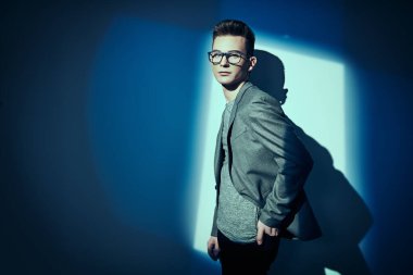Erkek güzelliği, moda. Klasik takım elbiseli ve gözlüklü yakışıklı genç adam stüdyoda mavi ışıkta beyaz duvarın yanında poz veriyor. Eğitim, eğitim..
