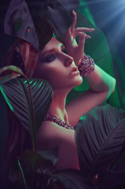 Kafasında boncuk ve eşarp takmış, tropikal bitkilerin arasında duran zarif bir kadının portresi. Güzellik, moda ve kozmetik konsepti. Tropikal ülkelerde tatil.