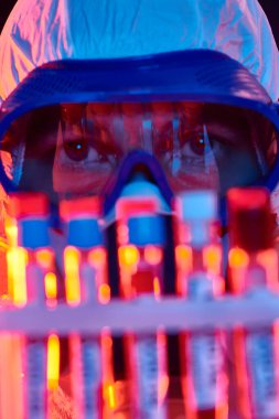 COVID-19. Koruyucu kıyafetli bir doktorun yakından çekilmiş portresi test tüplerinde enfekte olmuş insanların kan örneklerini gösteriyor. Coronavirus testi. Kırmızı ve mavi ışıklı siyah arkaplan.