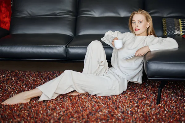Beautiful happy girl having a rest in her cozy living room, she is sitting on a soft carpet next to the sofa and drinks coffee. Home interior, furniture. Lifestyle.