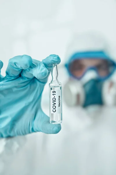 COVID-19 or SARS-CoV-2. Close-up portrait of a doctor microbiologist scientist in a protective PPE suit showing the ampoule with a vaccine against coronavirus. Vaccination.