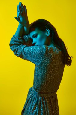 Lüks bir yaşam tarzı. Sarı arka planda parlak yeşil bir ışıkta parlak bir elbiseyle altın makyajla poz veren büyüleyici genç bir kadın. Parti biçimi.