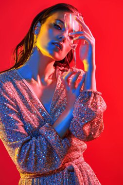 Makyaj ve makyaj malzemeleri. Renkli kırmızı, mavi ve sarı ışıkta elinde şampanya bardağıyla poz veren altın makyajlı genç bir kadının portresi. Parti biçimi. 