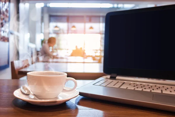 カフェの木製テーブルにラップトップでホット コーヒーの白いカップ. — ストック写真