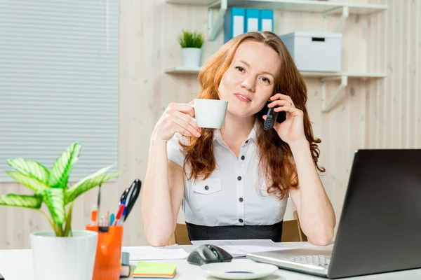 Mulher no escritório no trabalho com uma xícara de café e um telefone — Fotografia de Stock