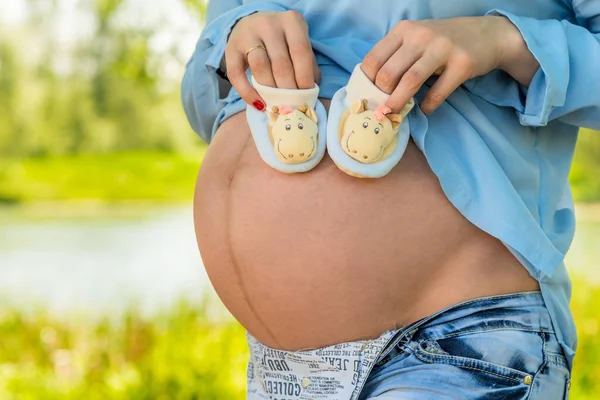 Vientre de una mujer embarazada primer plano y botines en las manos —  Fotos de Stock