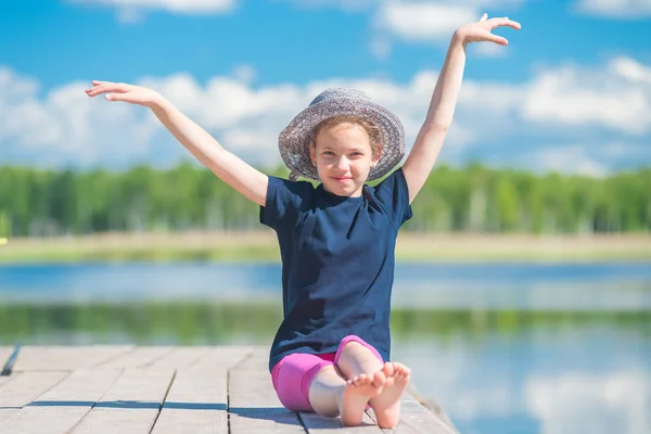 Meisje in een hoed zit op een houten pier in de buurt van een meer en poses — Stockfoto