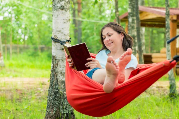 美丽的女人读一本书坐在一个红色的吊床在 g — 图库照片