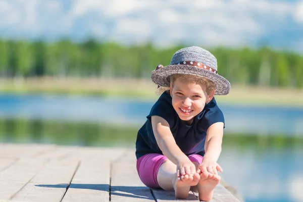 Gelukkig meisje in een hoed zit op een houten pier in de buurt van het meer en de pose — Stockfoto