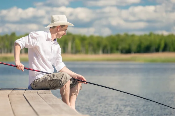 Pescador do sexo masculino com uma vara de pesca senta-se no cais perto do lago — Fotografia de Stock