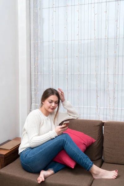 Νεαρή γυναίκα κάθεται στον καναπέ και χρησιμοποιώντας κινητό smartphone στην ορε — Φωτογραφία Αρχείου