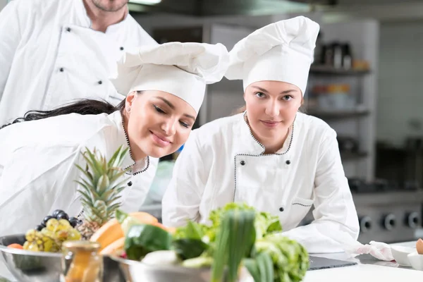 Dos jóvenes cocineras alegres usando uniforme trabajando en la cocina . — Foto de Stock