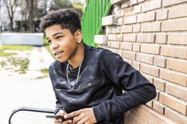 Genç afro çocuk bankta oturmuş akıllı telefonuyla tuğla duvarın arkasında sohbet ediyor ve gülümsüyor.