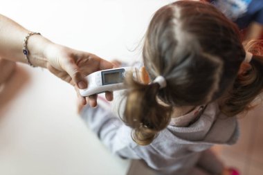 Tanınmayan bir anne, alnında bir termometreyle tanınmayan küçük kızının ateşini ölçer.