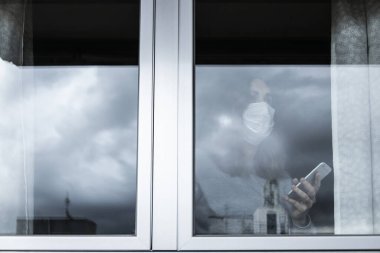 Solunum cihazı ve akıllı telefonu olan bir kadın belirsizlikle evinin penceresinden dışarı bakıyor ve karantinanın sona ermesini bekliyor.