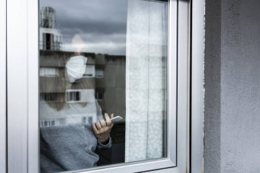 Solunum cihazı ve akıllı telefonu olan bir kadın belirsizlikle evinin penceresinden dışarı bakıyor ve karantinanın sona ermesini bekliyor.