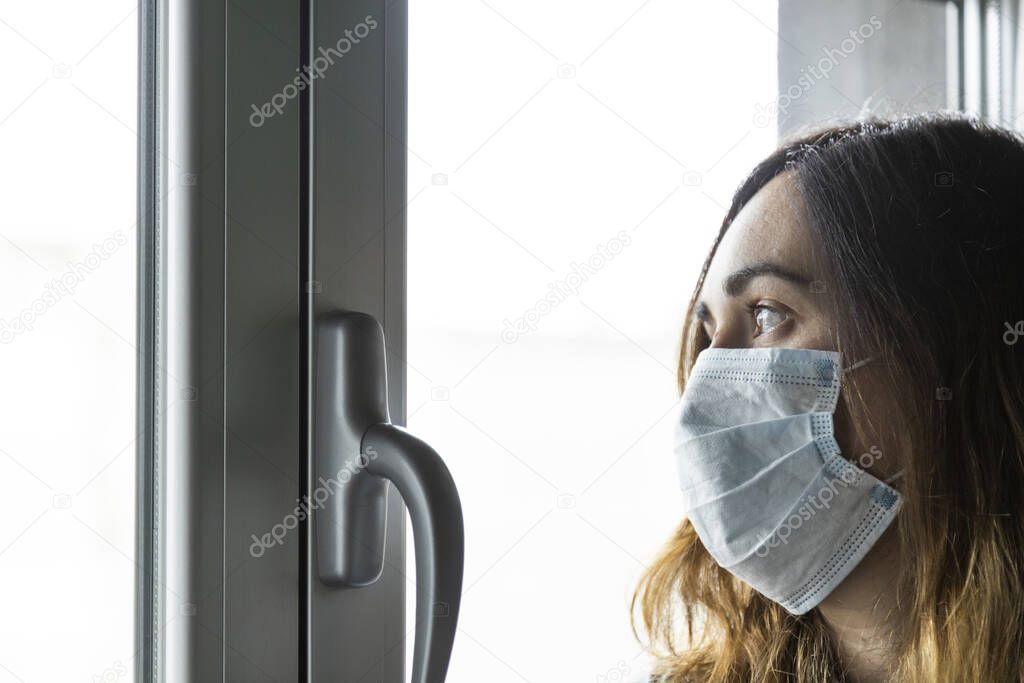 woman with respirator looking through the window of her house with uncertainty, waiting for the quarantine to end by covid-19