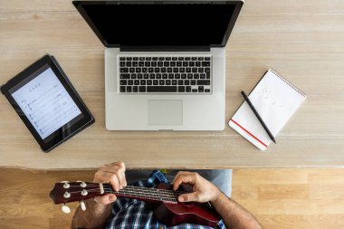 Tanınmayacak halde ukulelesini evde dizüstü bilgisayar ve tabletle prova eden adam.