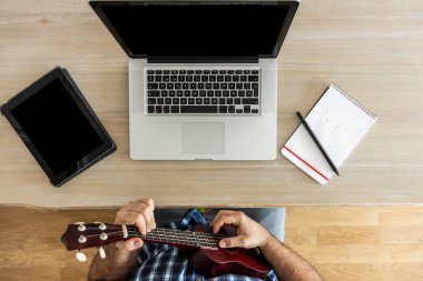 Tanınmayacak halde ukulelesini evde dizüstü bilgisayar ve tabletle prova eden adam.