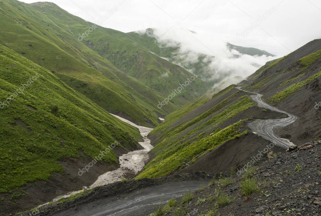 dangerous gravel road and track of the kaldamo pass with green mountains with snow and foggy clouds in the valley, Kyrgyzstan.