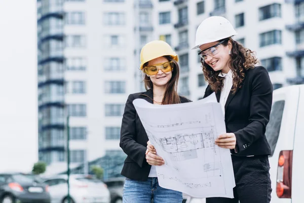 Two business women in building helmets and safety glasses looking at building schemes, architectural concept