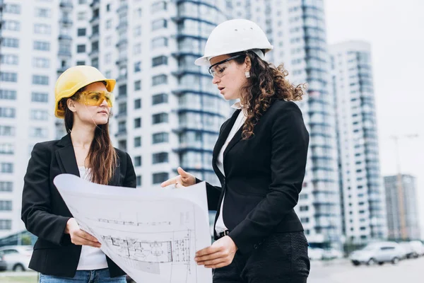 Two business women in building helmets and safety glasses looking at building schemes, architectural concept