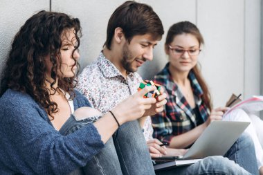Genç insanlar farklı araçları kullanma ve gülümseyerek, duvara yakın oturuyor. Dizüstü bilgisayar kullanan okuyan öğrenciler. Eğitim sosyal medya kavramı.