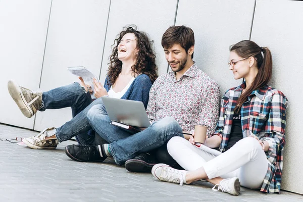 Mladí lidé jsou pomocí různých miniaplikací a usmíval se, seděl u zdi. Studenti studující pomocí přenosného počítače. Koncepce vzdělávání sociální média. — Stock fotografie