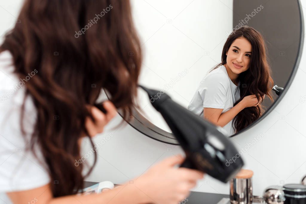 Beautiful young woman wearing a white t-shirt, looking at the mirror drying her hair smiling, windy hair. Beauty concept photo, lifestyle