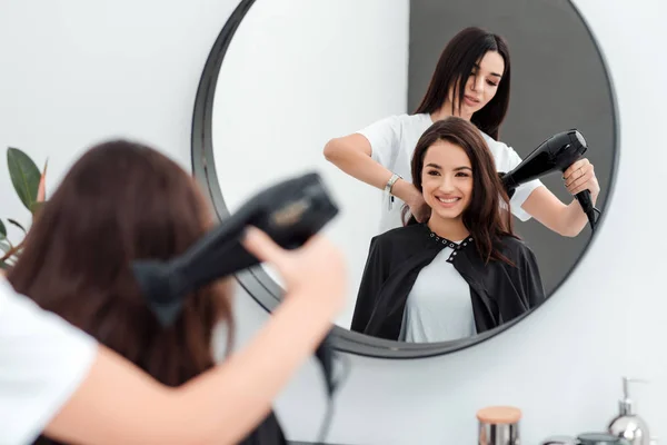 Милая красивая девушка с длинными волосами смотрит в зеркало, она пришла в салон красоты, ее парикмахер работает с ее волосами . — стоковое фото