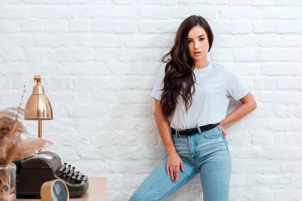 Stylish girl dressed in casual style, standing against a background of brick, white wall around her on the table stands a printing machine and a lamp — 图库照片