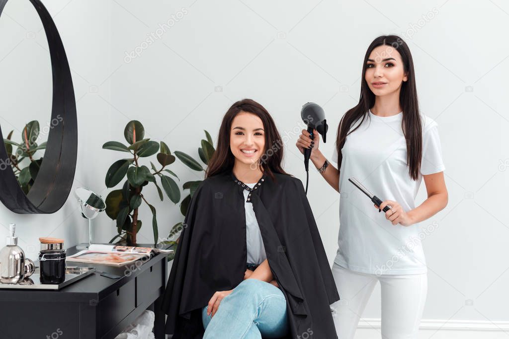 Smiling client sits in hairdresser's chair, hairdresser standing beside her with drying and comb in her hands