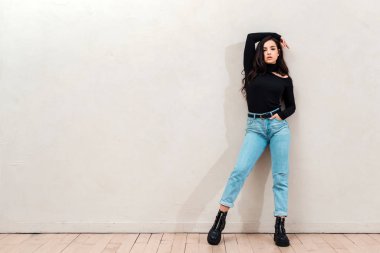 Beautiful brunette wearing a black cloth, throws her hand over her head and poses against a beige wall background. Fashion concept, place for advertising, copyspace