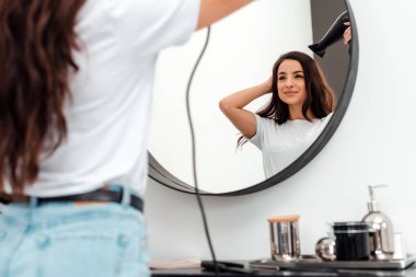 Pretty young woman dries her beautiful hair and looks in the mirror
