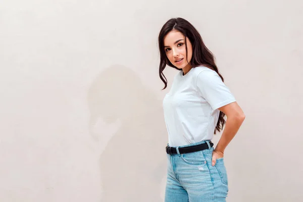 Attractive brunette put her hands in her back pockets, cute posing against a beige wall background — Stock Photo, Image